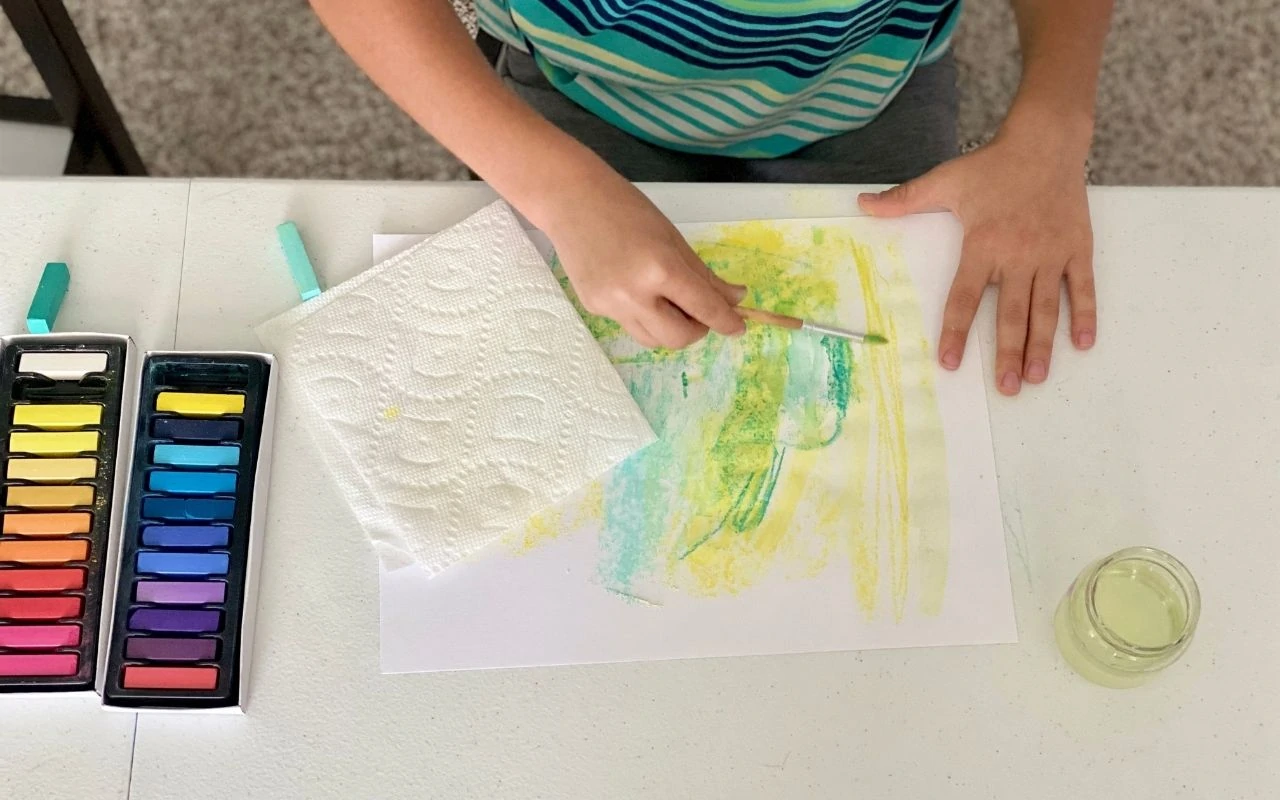 Child makes marks with chalk pastel and uses paint brush and water over it to fill paper
