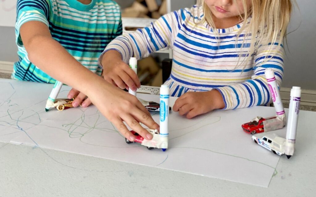 Child driving toy car across white paper with marker attached by tape to it
