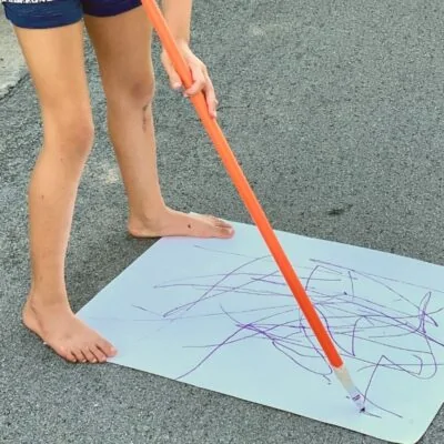 Child holding a long stick with a marker attached to the bottom to draw while standing up
