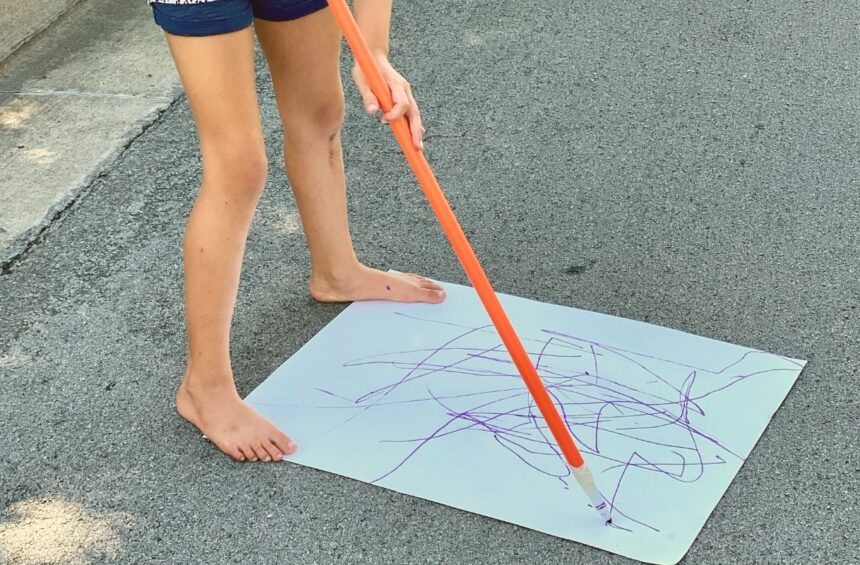 Child holding a long stick with a marker attached to the bottom to draw while standing up