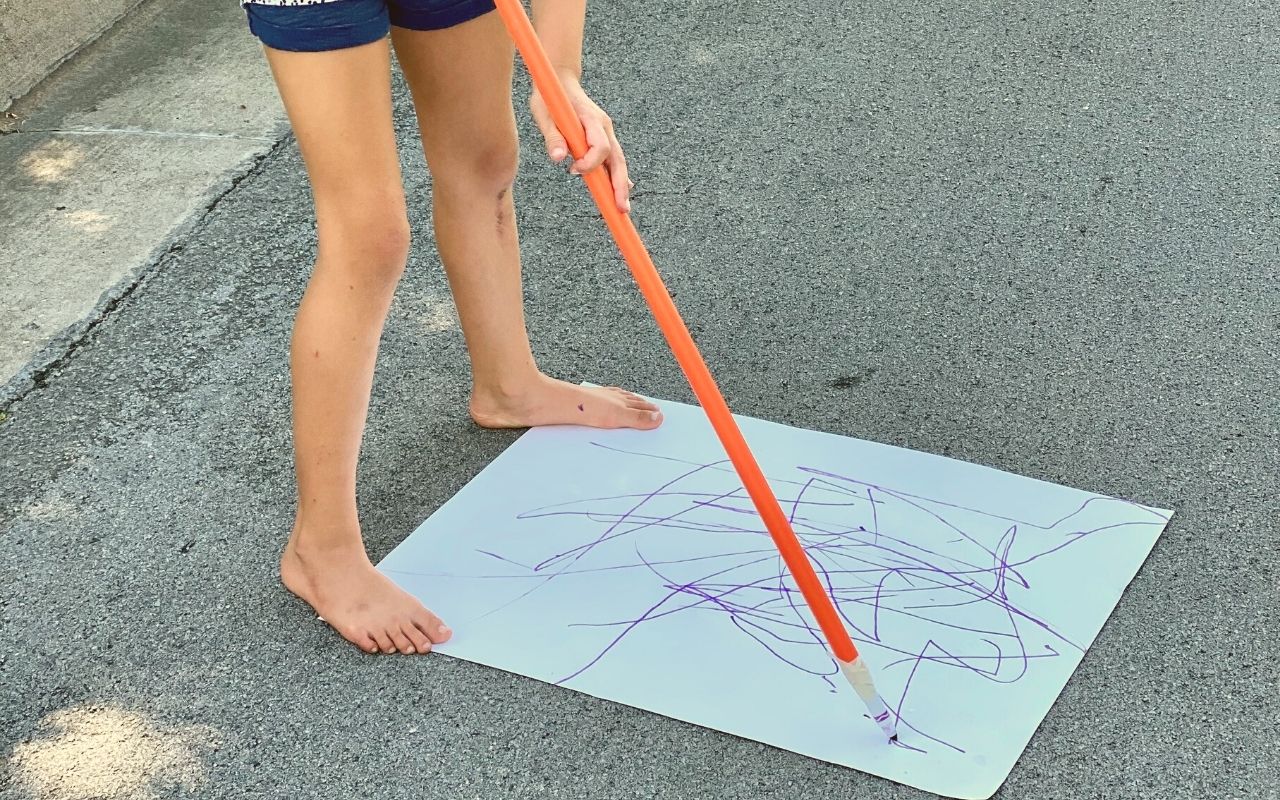 Child holding a long stick with a marker attached to the bottom to draw while standing up