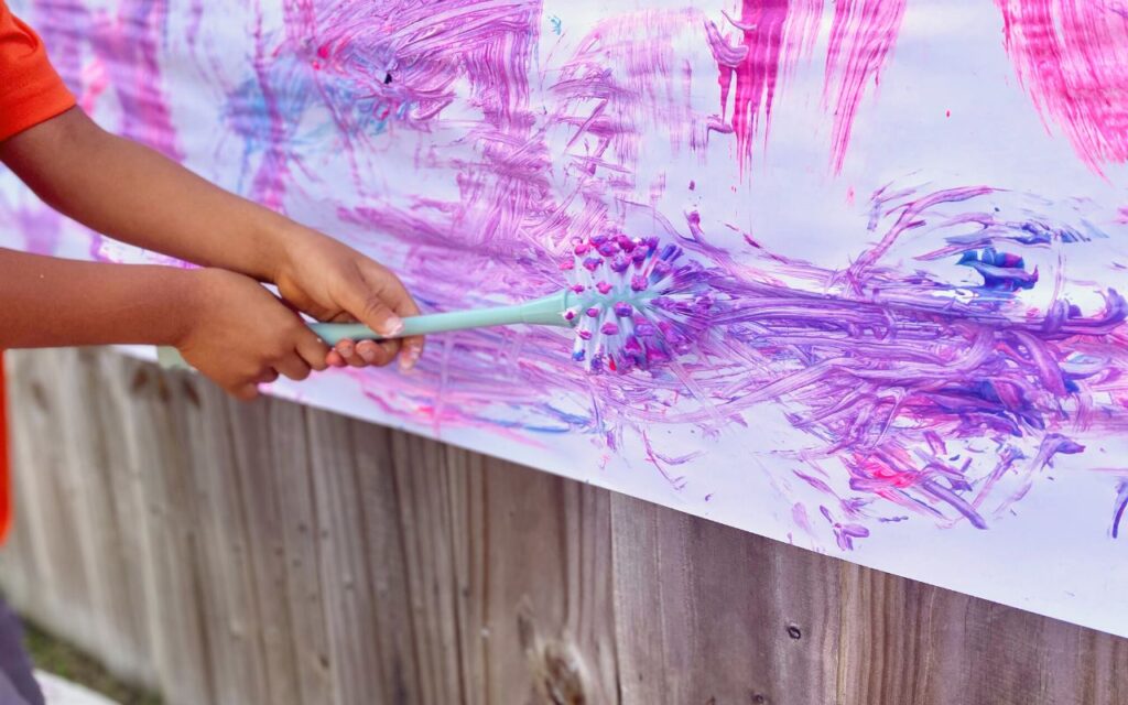Child holding cleaning brush while painting on large hanging paper