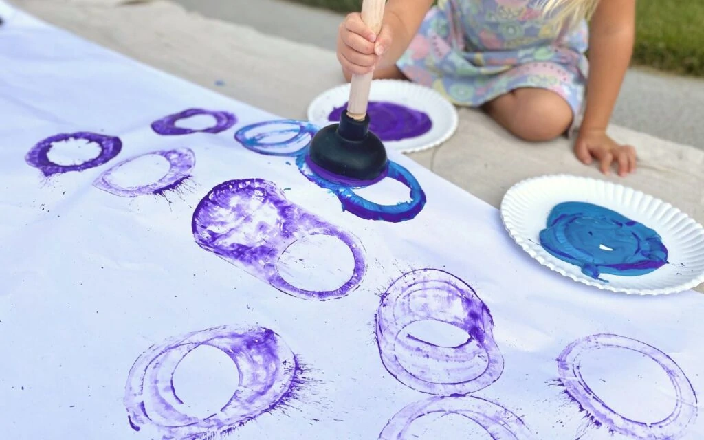 Child makes paint marks on large paper using a plunger to paint