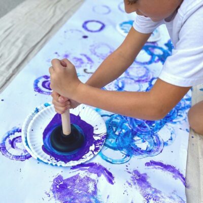 Child makes paint marks on large paper using a plunger to paint