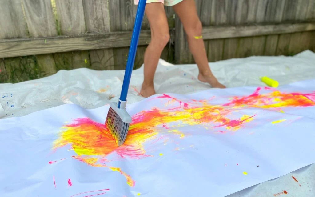 Child using long kitchen broom to swish paint around paper