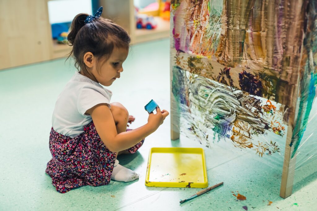 young child painting with a sponge, brushes and paints on a cling film wrapped all the way round a table